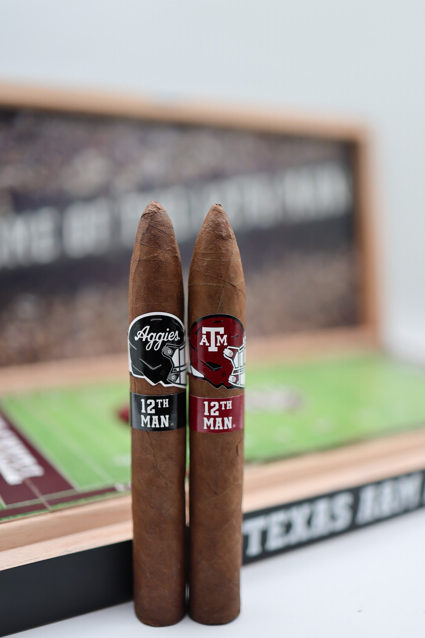 Two cigars with '12th Man' and 'Aggies' labels in front of a blurred sports-themed background.